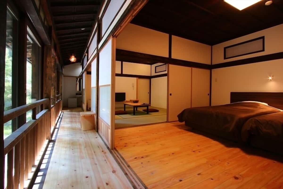 Una habitación tradicional japonesa con suelo de tatami, una cama baja de madera, puertas correderas y vistas a través de una ventana con marco de madera.