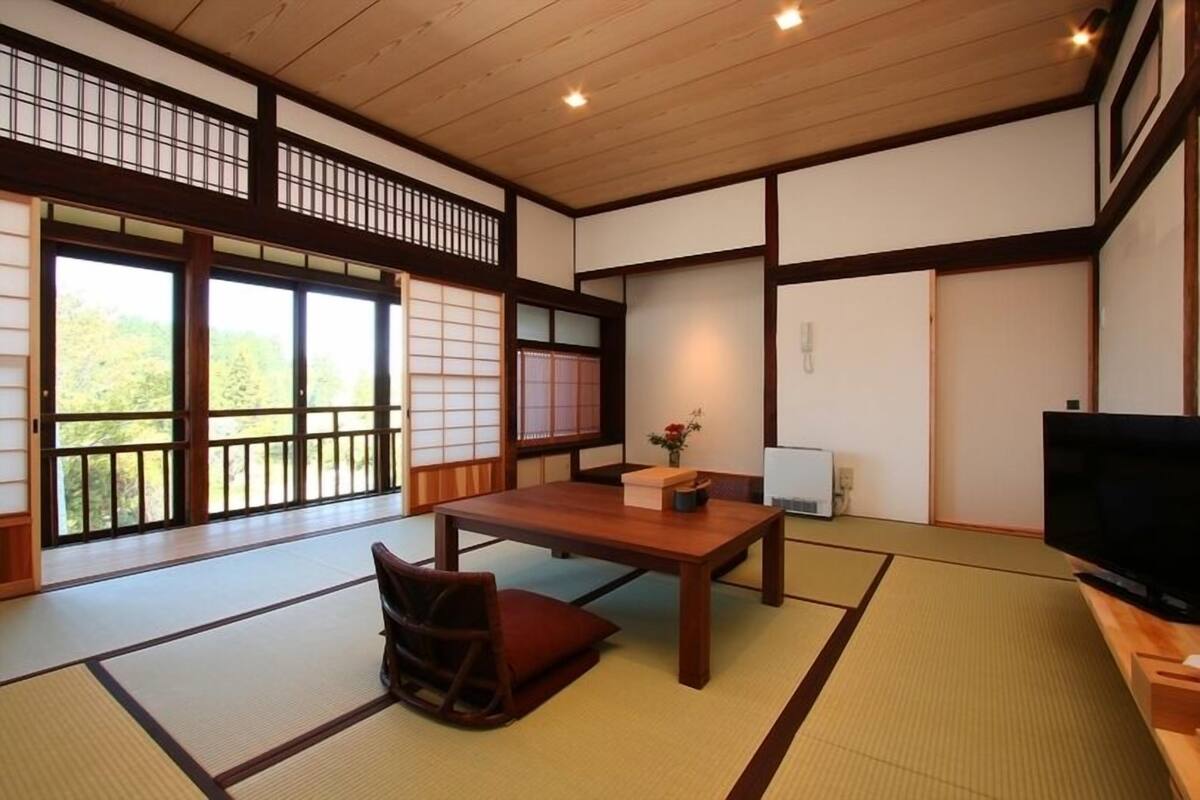 Una habitación japonesa tradicional con suelo de tatami, una mesa baja, una silla, un televisor y puertas correderas.