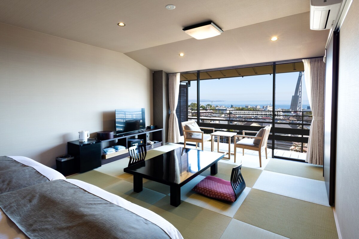 A hotel room with a bed, a desk, a chair, a TV, and a balcony with a city view.