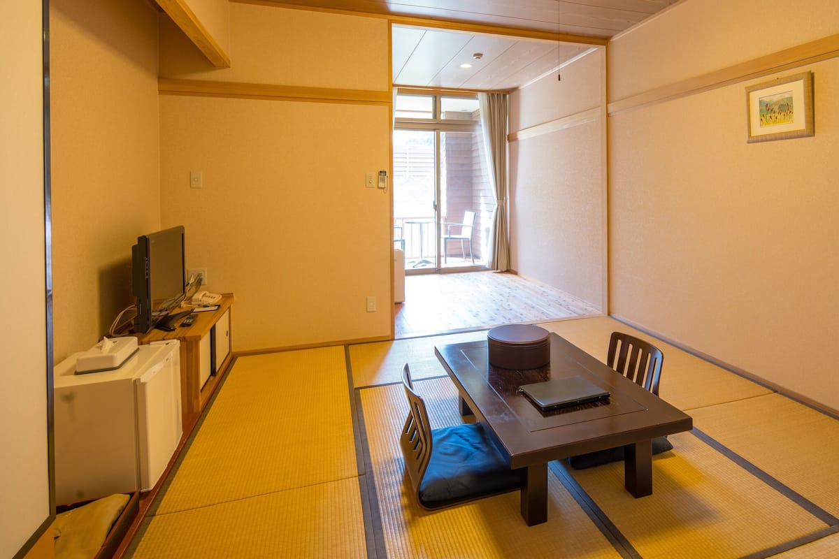 Una habitación japonesa tradicional con suelo de tatami, una mesa baja con cojín, un televisor y un pequeño escritorio con teléfono.