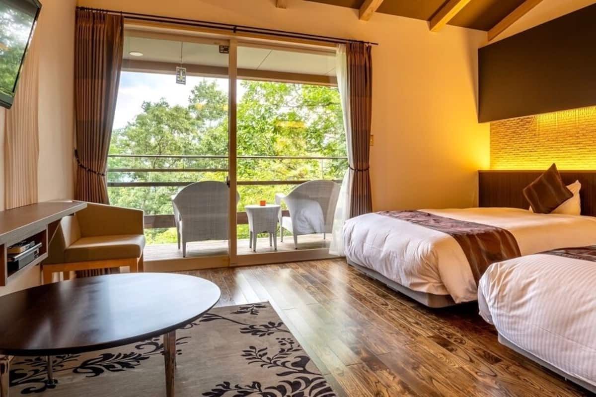 A hotel room with a bed, a desk, a chair, a small table, and a balcony with a view of trees.