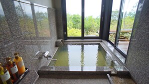 A modern bathroom with a large bathtub, a window with a view of greenery, and a shelf with toiletries.