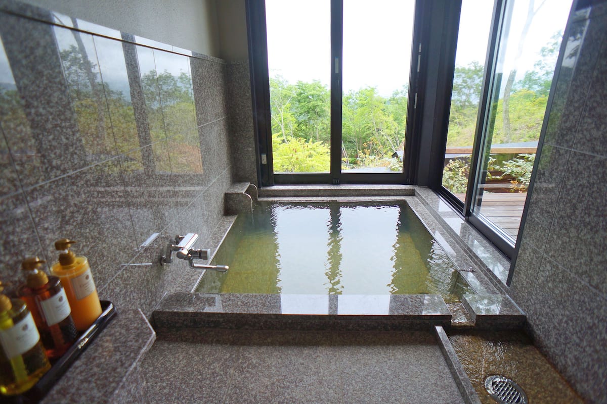 A modern bathroom with a large bathtub, a window with a view of greenery, and a shelf with toiletries.
