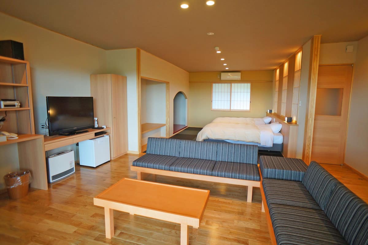 A modern hotel room with a bed, a TV, a desk, a small fridge, and a seating area with a sofa and a bench.