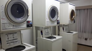 A row of front-loading washing machines in a laundry room with instructional labels and a window with curtains. - Hotel Crown Hills Sagamihara (Sagamihara)