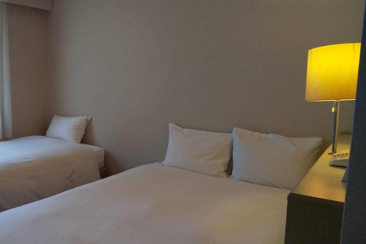 A hotel room with two beds, a bedside table with a lamp, and a window with sheer curtains.