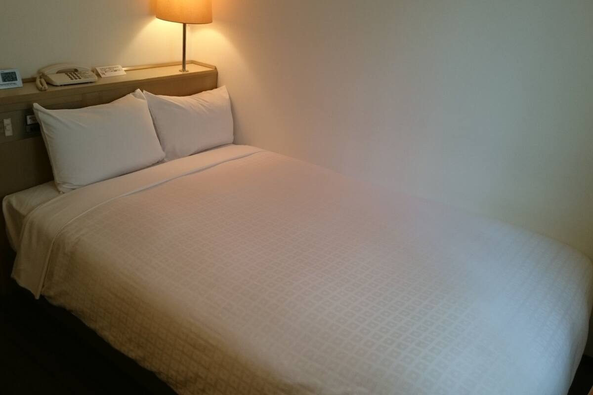 A hotel room with a bed, a nightstand with a lamp, a phone, and a clock.