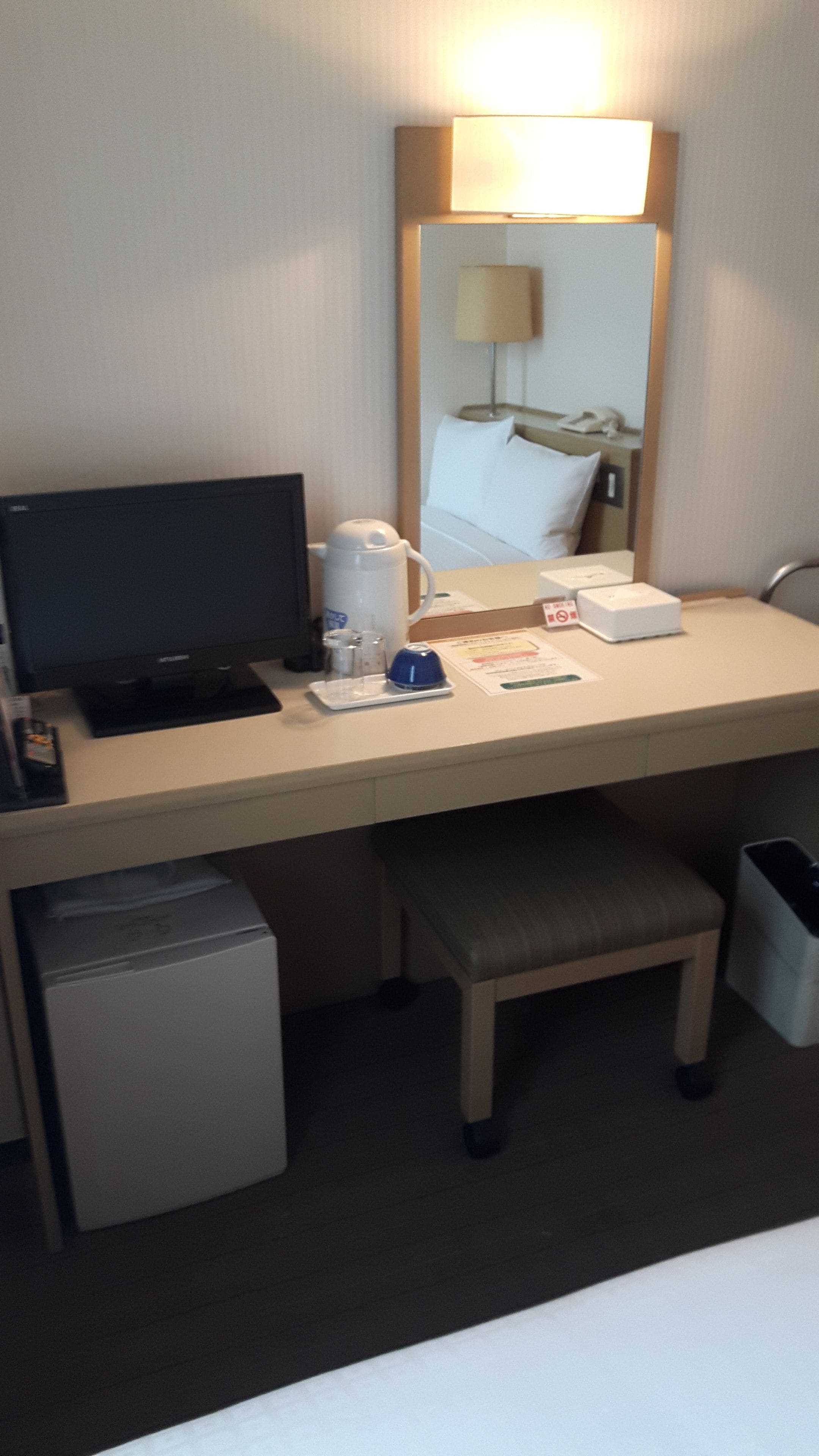 A hotel room with a desk, a television, a coffee maker, a mini-fridge, and a trash bin.
