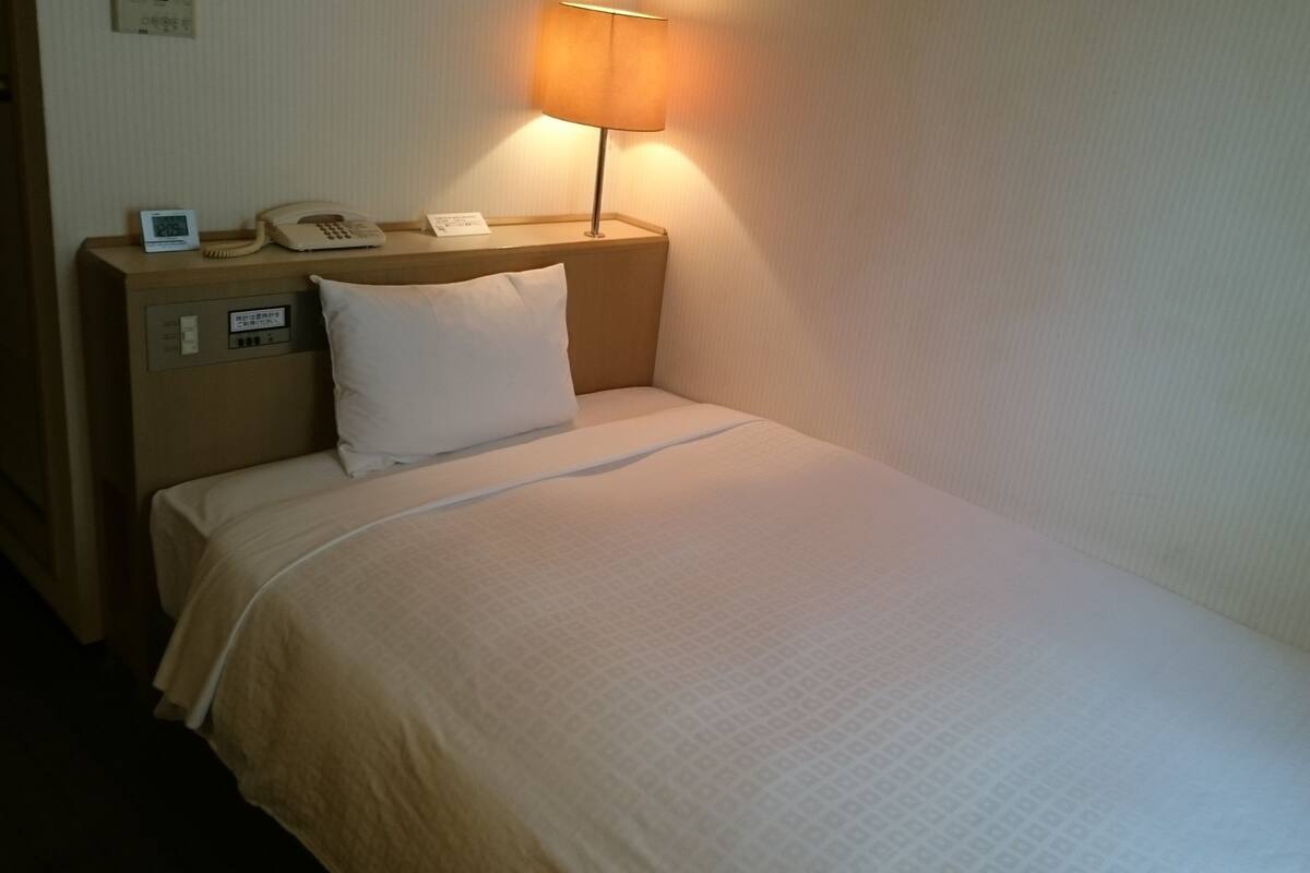 A hotel room with a single bed, a bedside table with a lamp, a phone, and a clock.