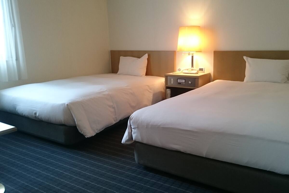 A hotel room with two beds, a nightstand with a lamp, and a window with curtains.