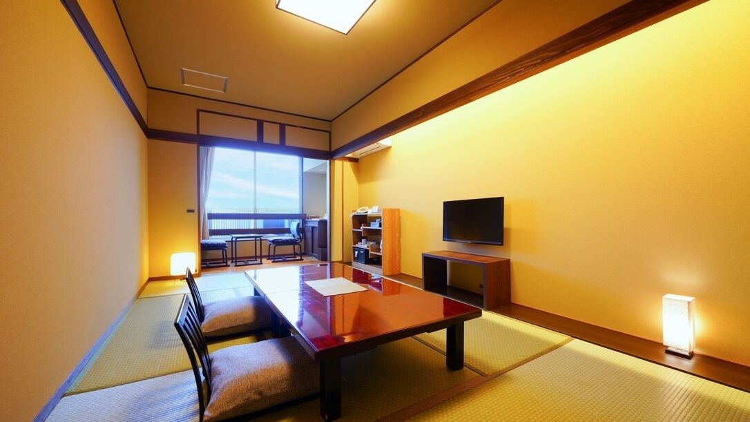 Ein traditioneller japanischer Raum mit Tatami-Böden, einem niedrigen Tisch, Kissen, einem Fernseher und Blick ins Freie.
