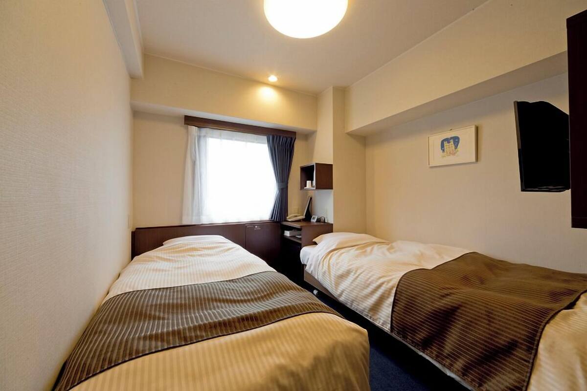 A hotel room with two beds, a window with curtains, a bedside table with a lamp, and a wall-mounted TV.