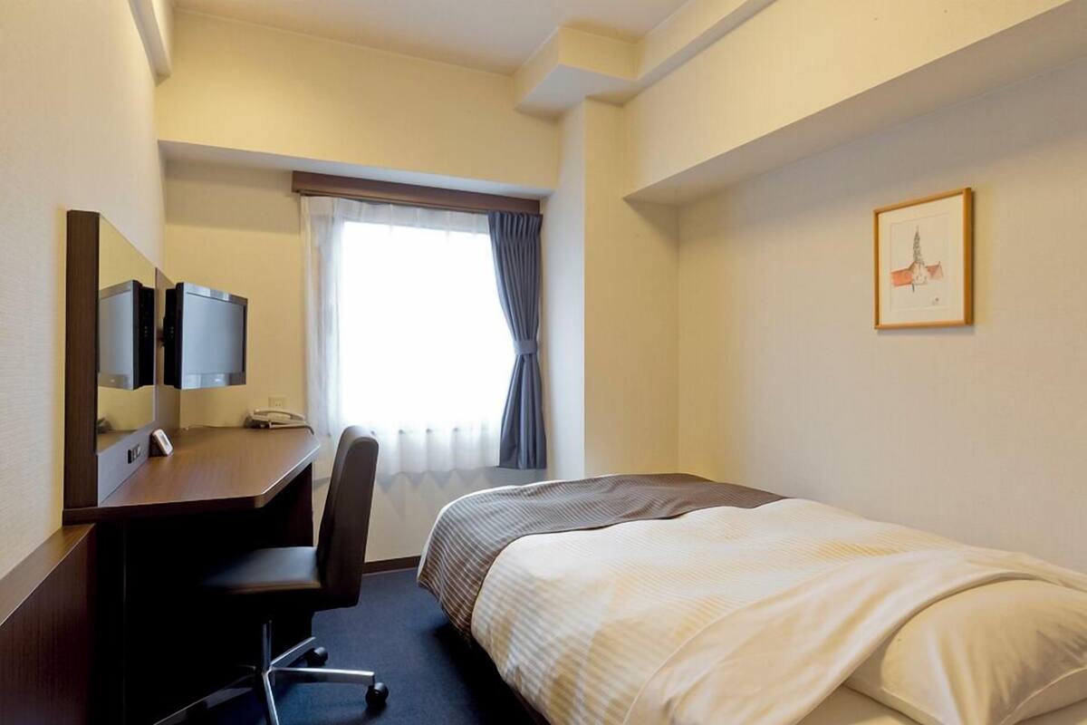 A hotel room with a bed, a desk with a chair, a television, and a window with curtains.