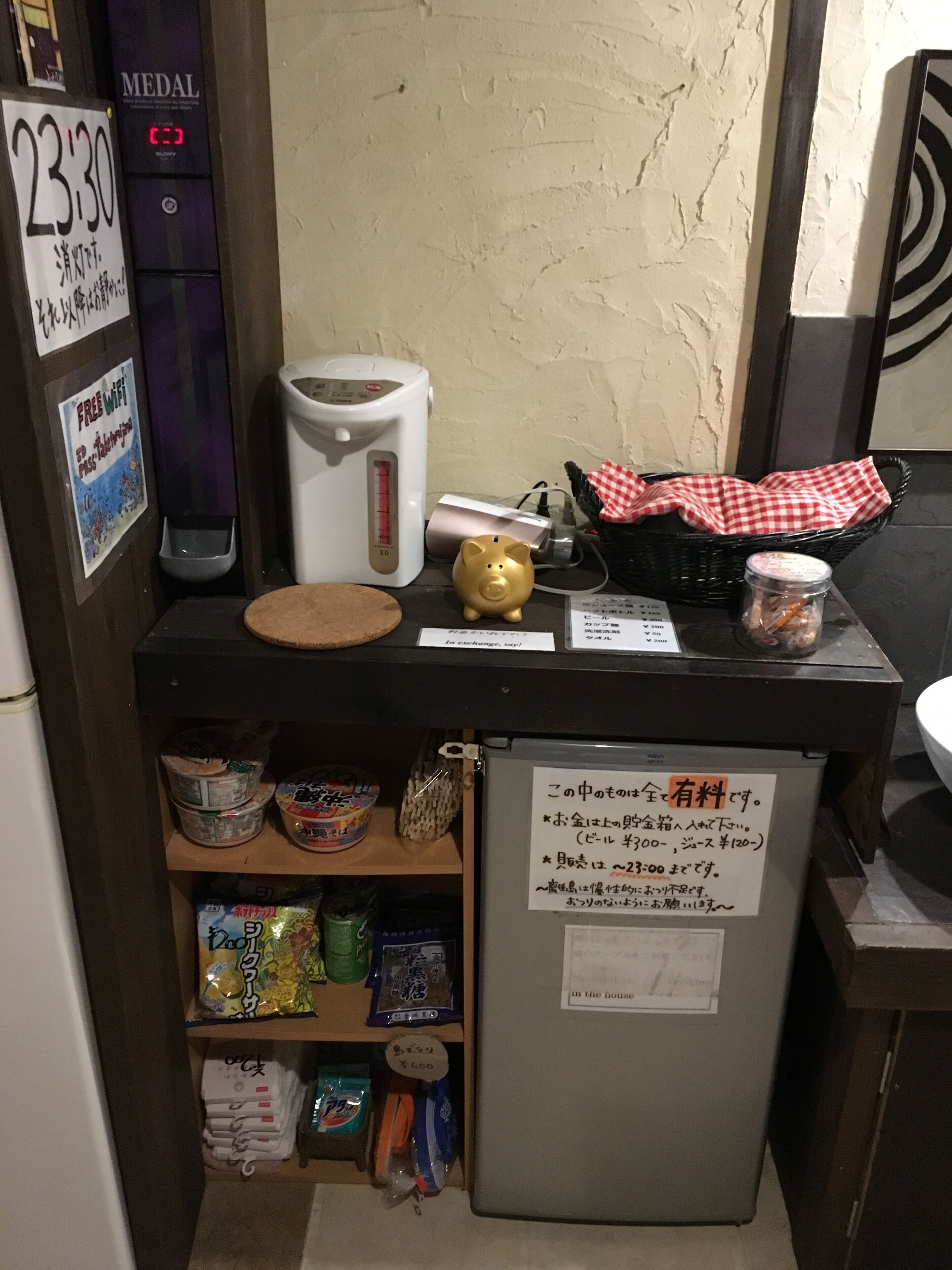 Une petite cuisine équipée d’un four à micro-ondes, d’un cuiseur à riz, d’une caisse enregistreuse et de divers produits alimentaires rangés sur des étagères.