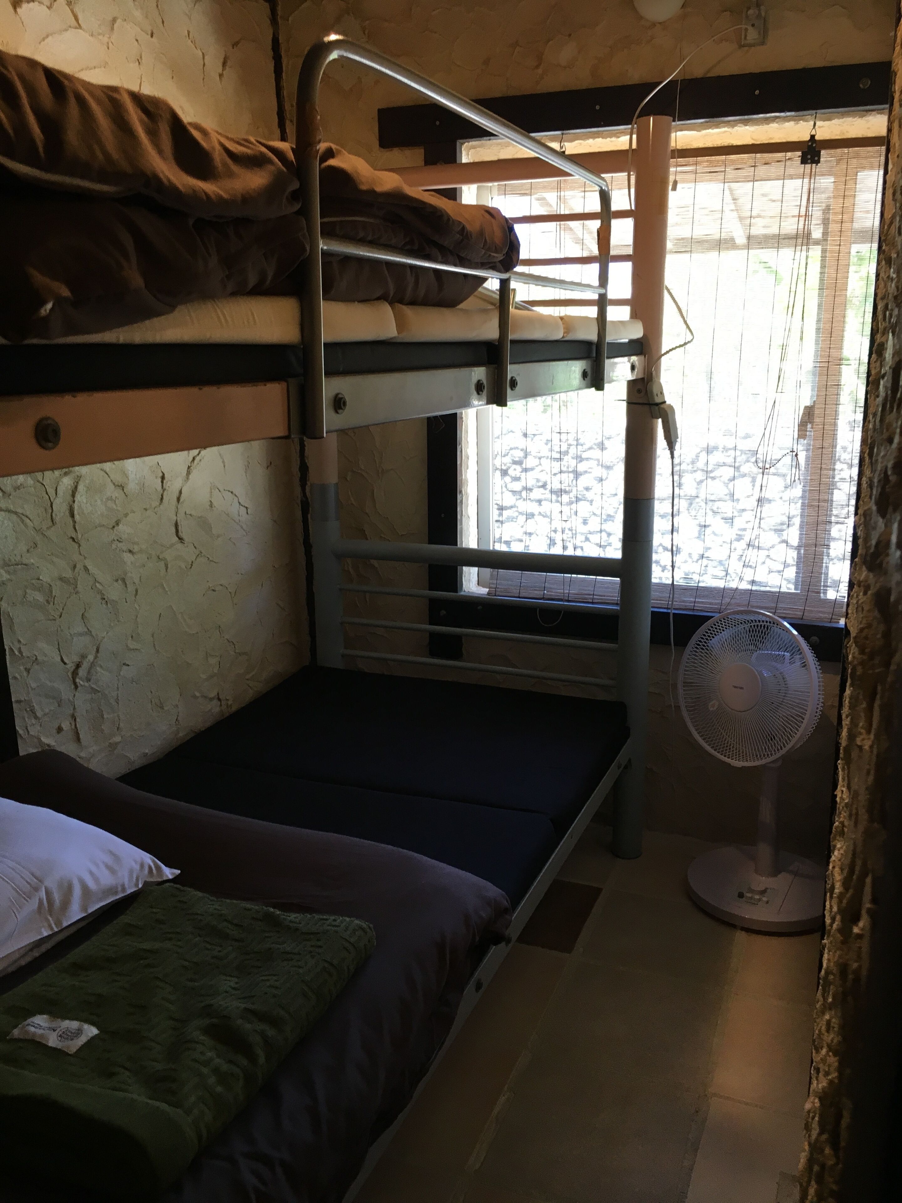 A bunk bed room with a window, a standing fan, and a green blanket on the lower bunk.