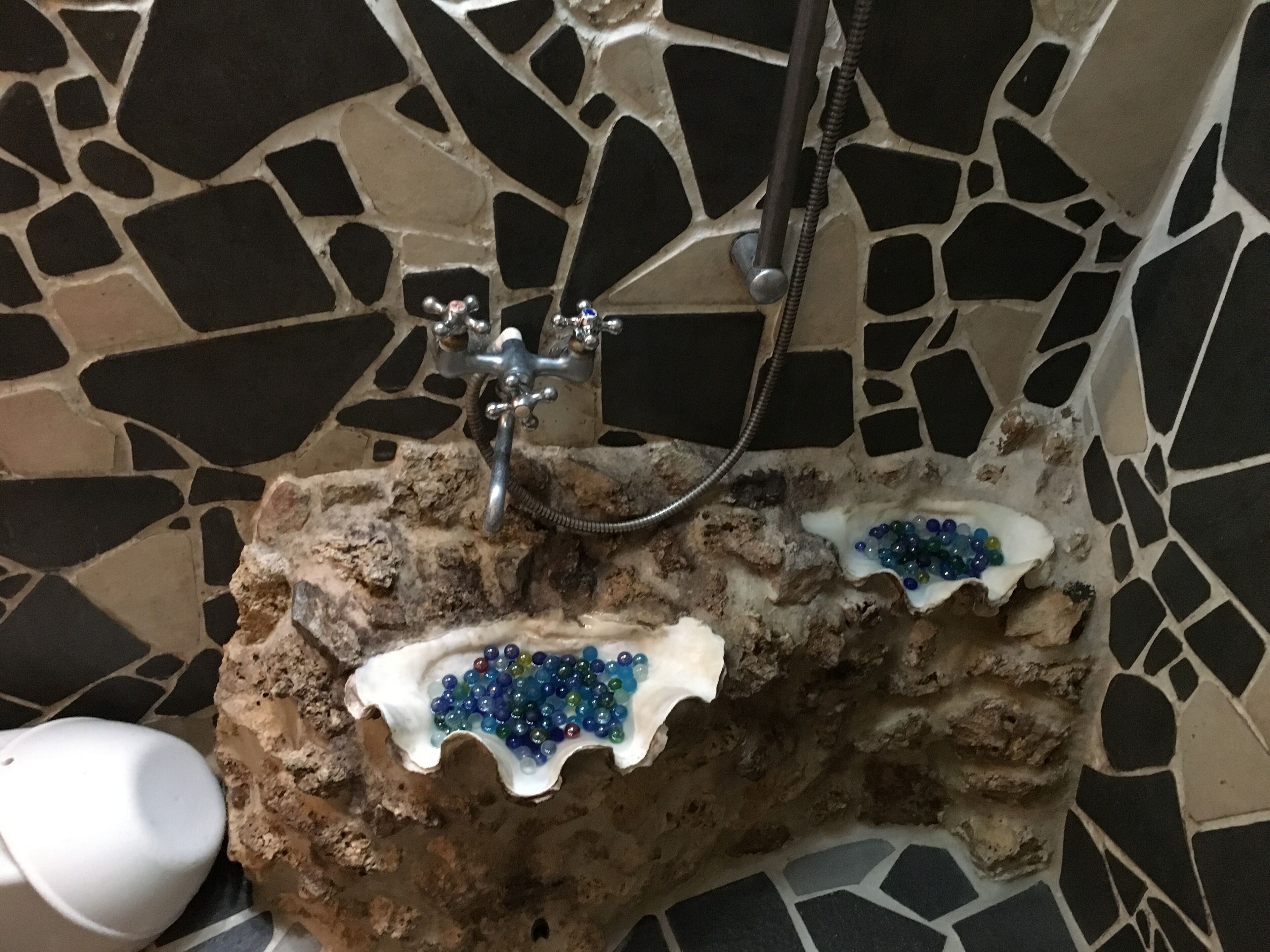 Une salle de bain avec un mur recouvert de mosaïques, un lavabo en pierre agrémenté de petits galets bleus et une douchette à main.