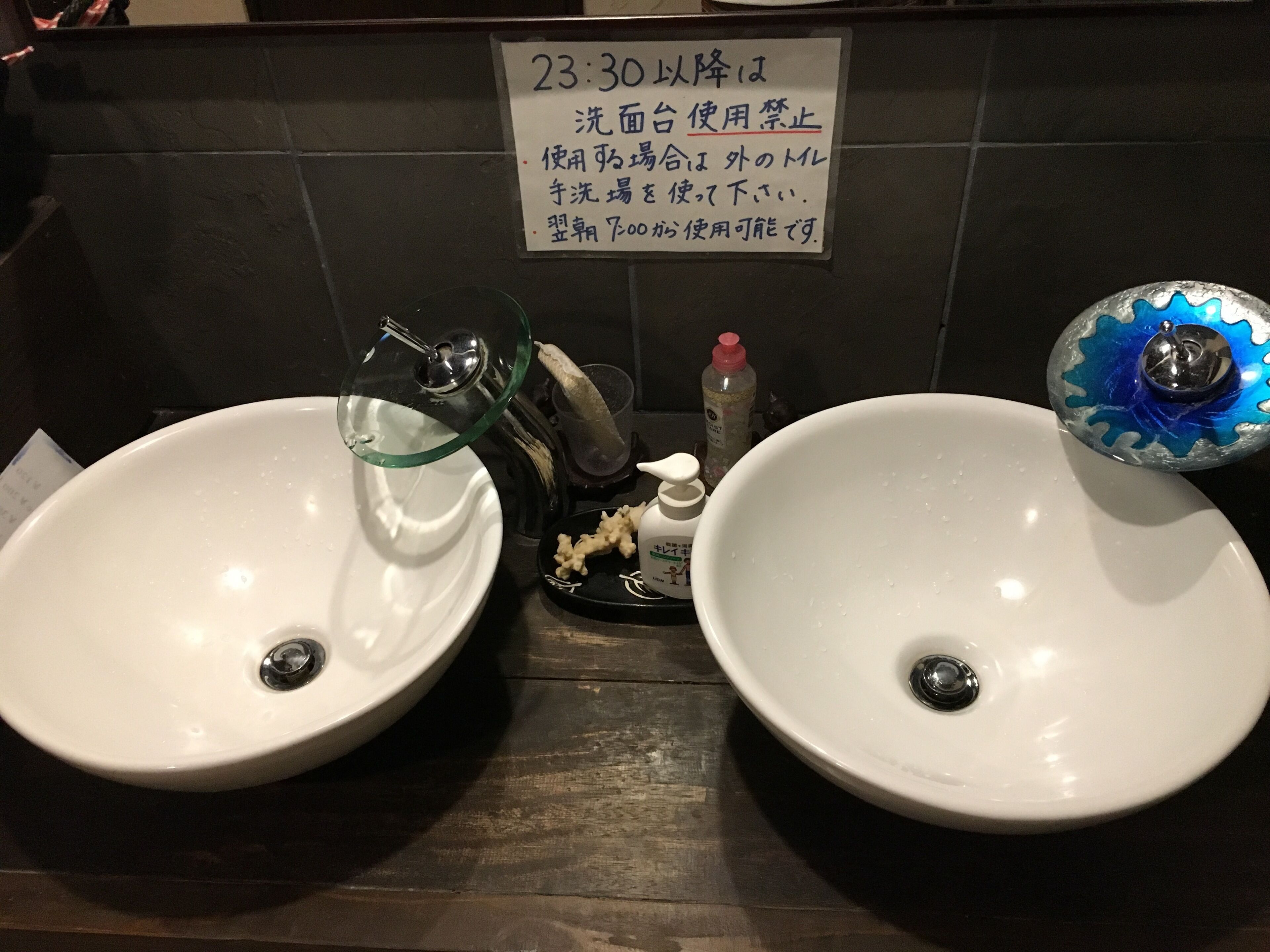 Two white sinks with glass covers, a soap dispenser, and a sign in Japanese.