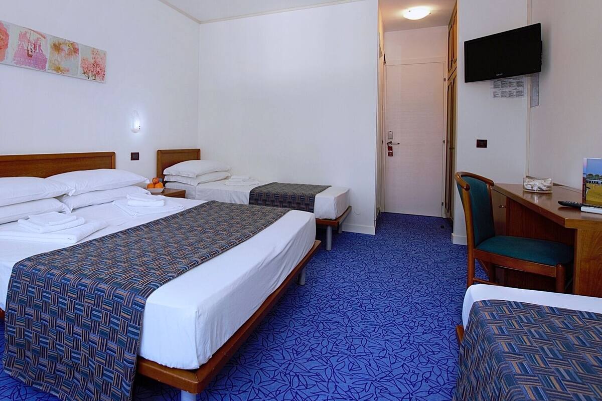 Quarto de hotel com duas camas, uma escrivaninha, uma cadeira, uma televisão e a porta do banheiro.