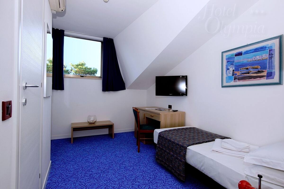 Quarto compacto com cama, escrivaninha, cadeira, televisão e janela com cortinas.