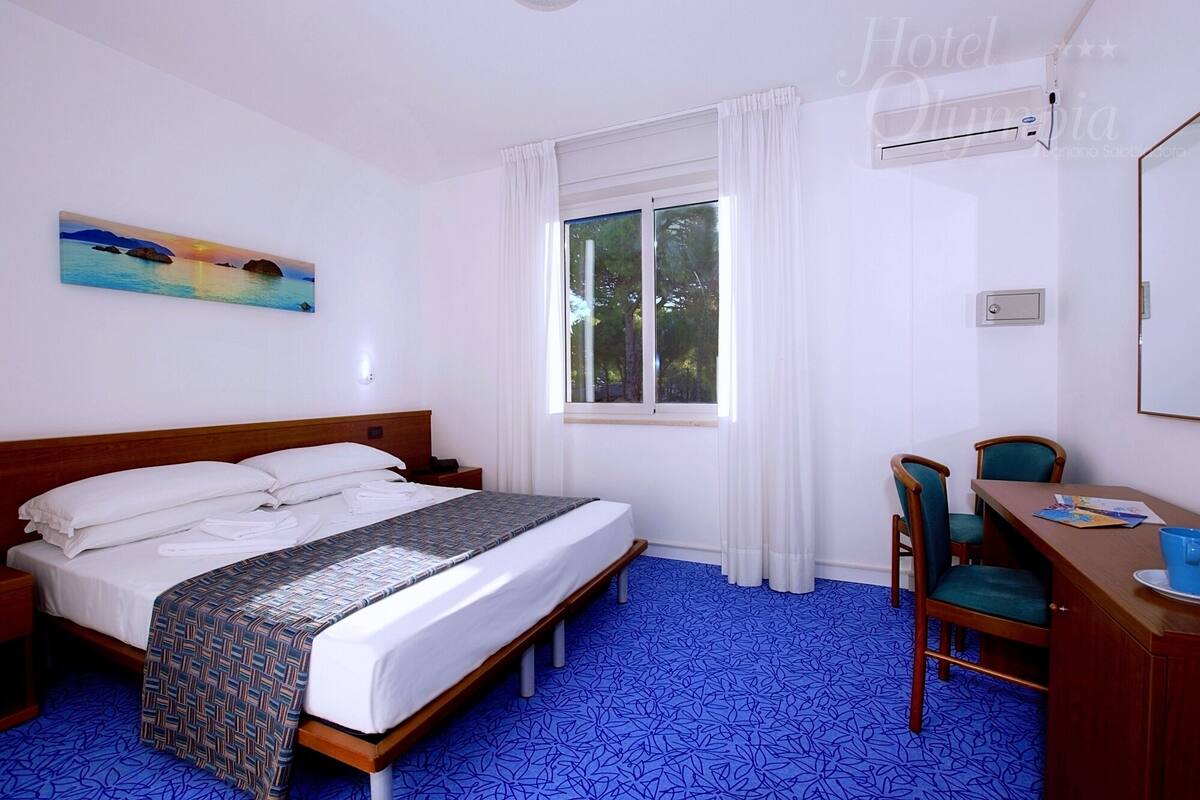 Quarto de hotel com cama, escrivaninha, cadeira, janela e um quadro na parede.