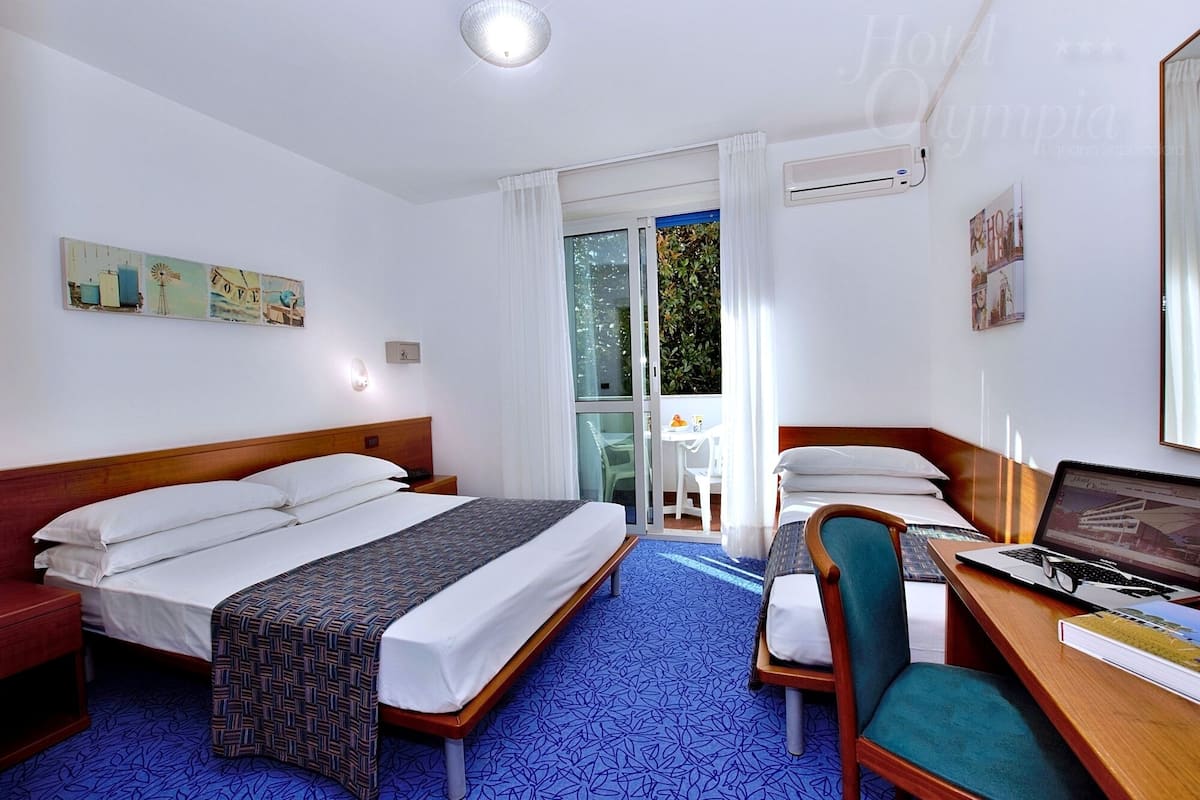 Quarto de hotel com duas camas, uma escrivaninha com um laptop, uma cadeira e uma janela grande com vista para vegetação.