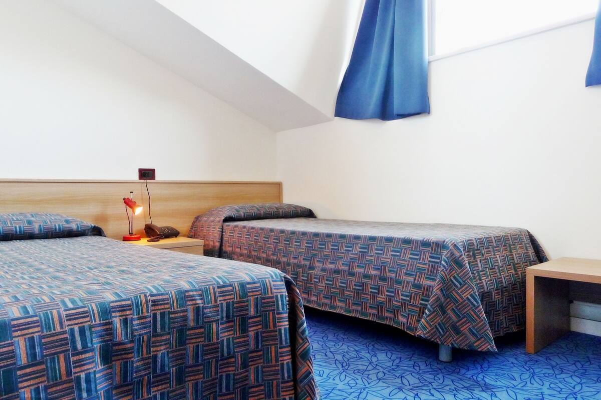 Quarto com duas camas, cabeceira de madeira, escrivaninha e janela com cortinas azuis.