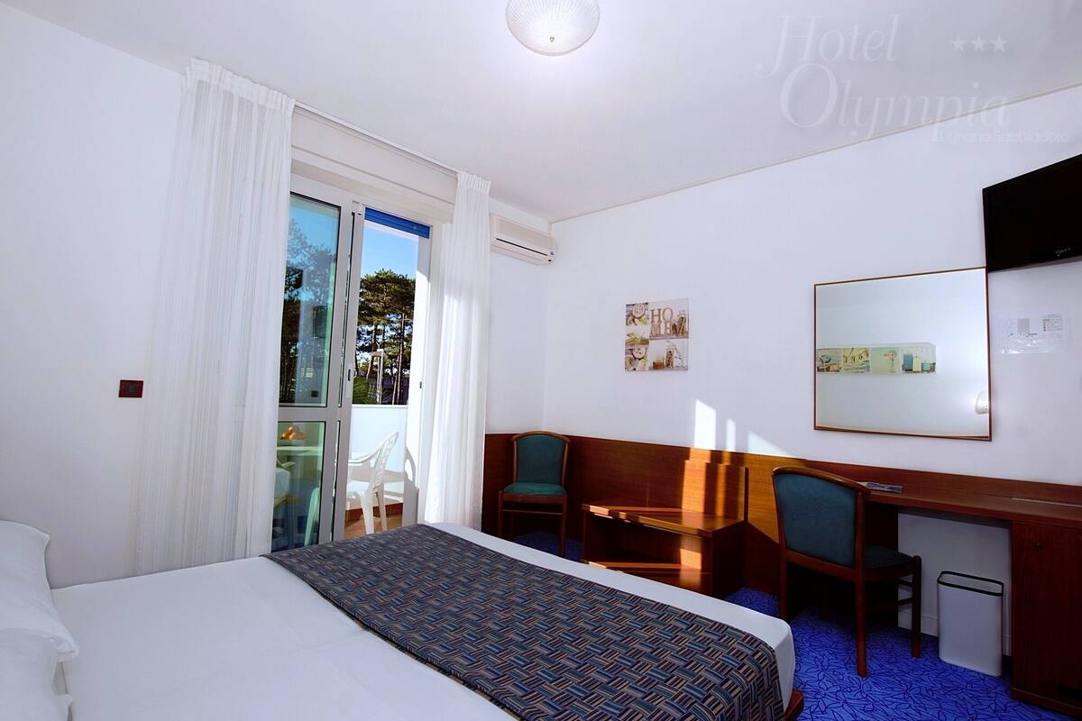 Quarto de hotel com uma cama, uma escrivaninha, duas cadeiras, uma mesinha e um espelho.