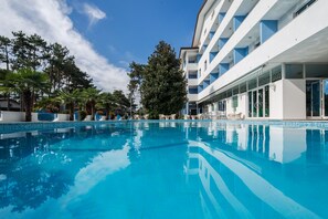 A swimming pool with clear blue water, surrounded by a modern building with balconies and a large tree in the center.
