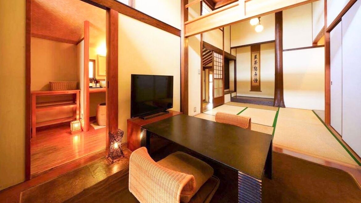 Una habitación japonesa tradicional con suelo de tatami, una televisión, una mesa baja y puertas correderas que dan a otra habitación.