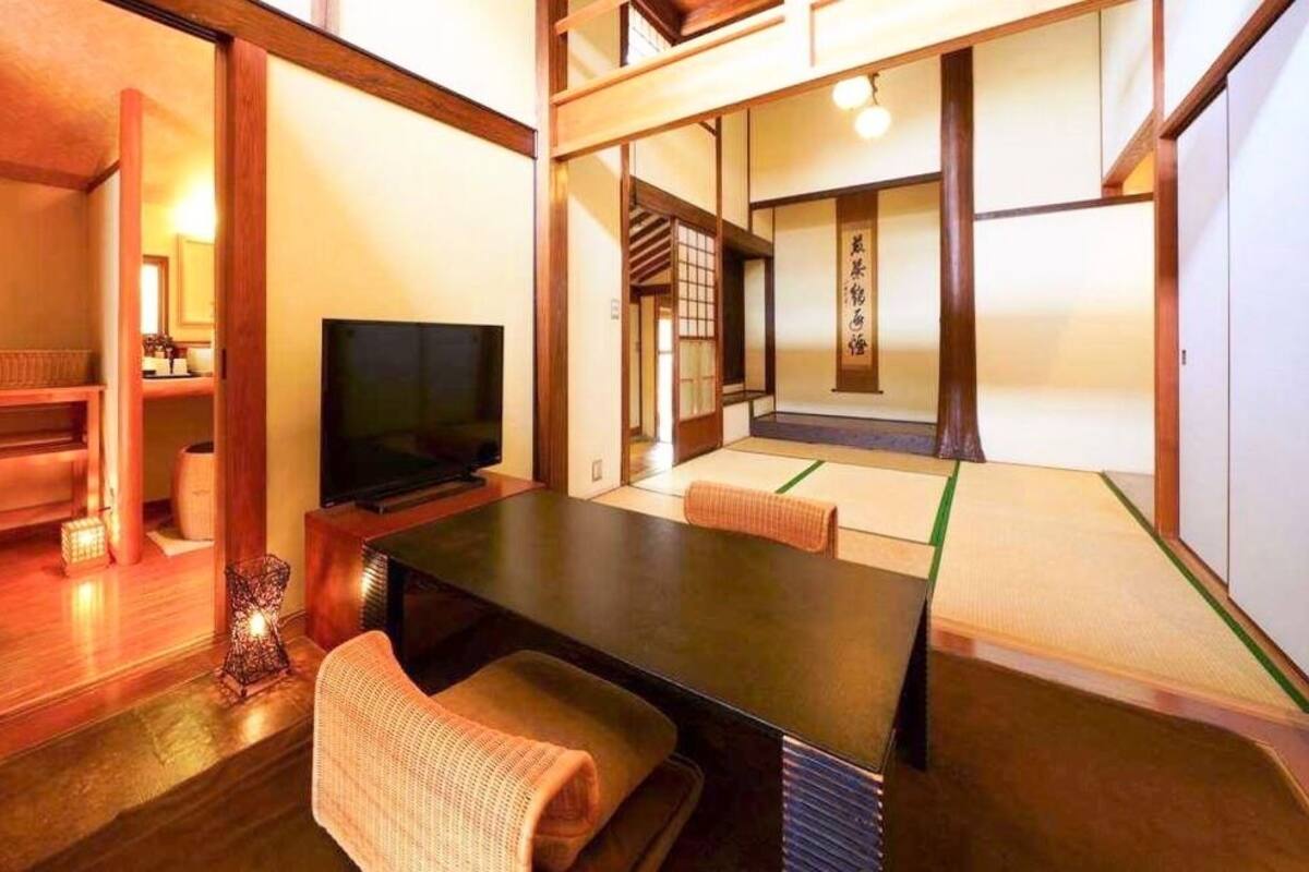Una habitación japonesa tradicional con suelo de tatami, una televisión, una mesa baja y puertas correderas que dan a otra habitación.