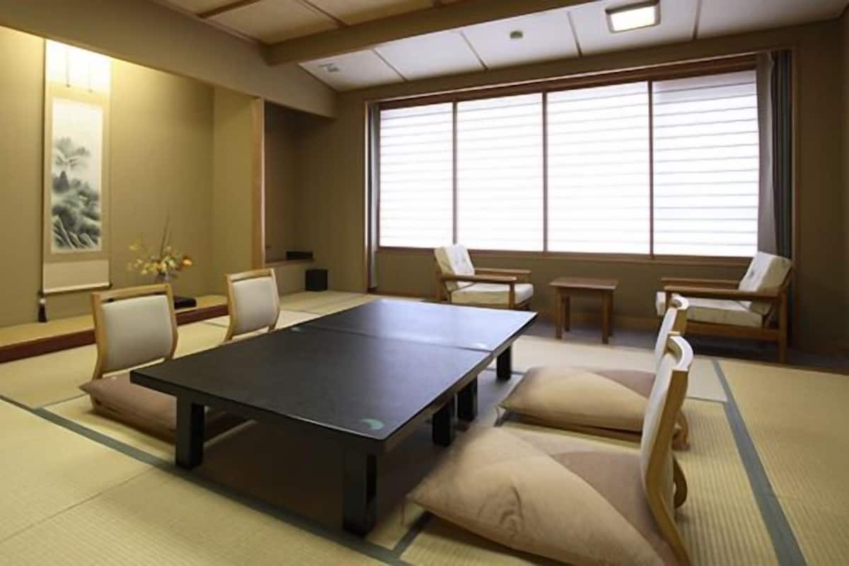 Une pièce traditionnelle avec un sol recouvert de tatamis, une petite table et des chaises, de grandes fenêtres équipées de stores et un tableau accroché au mur.