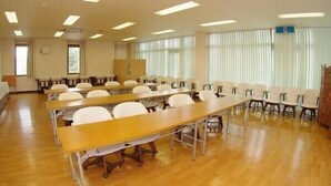 Une salle de conférence avec des rangées de tables et de chaises, de grandes fenêtres équipées de stores et un éclairage fluorescent au plafond.
