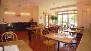 Un restaurant avec des tables et des chaises en bois, un comptoir avec une caisse enregistreuse et de grandes fenêtres laissant entrer la lumière naturelle.
