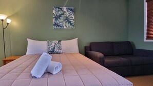 A neatly made bed with a quilted cover, a pair of rolled towels, a wall-mounted plant, a floor lamp, and a dark-colored sofa. - Canoe Safaris Lodge (Ohakune)