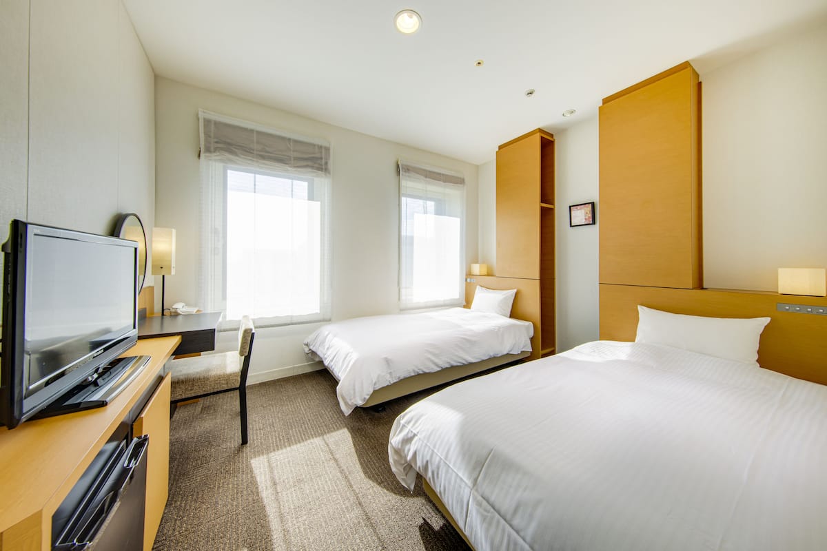 Quarto de hotel com duas camas de solteiro, uma televisão, uma escrivaninha com cadeira e uma janela grande.