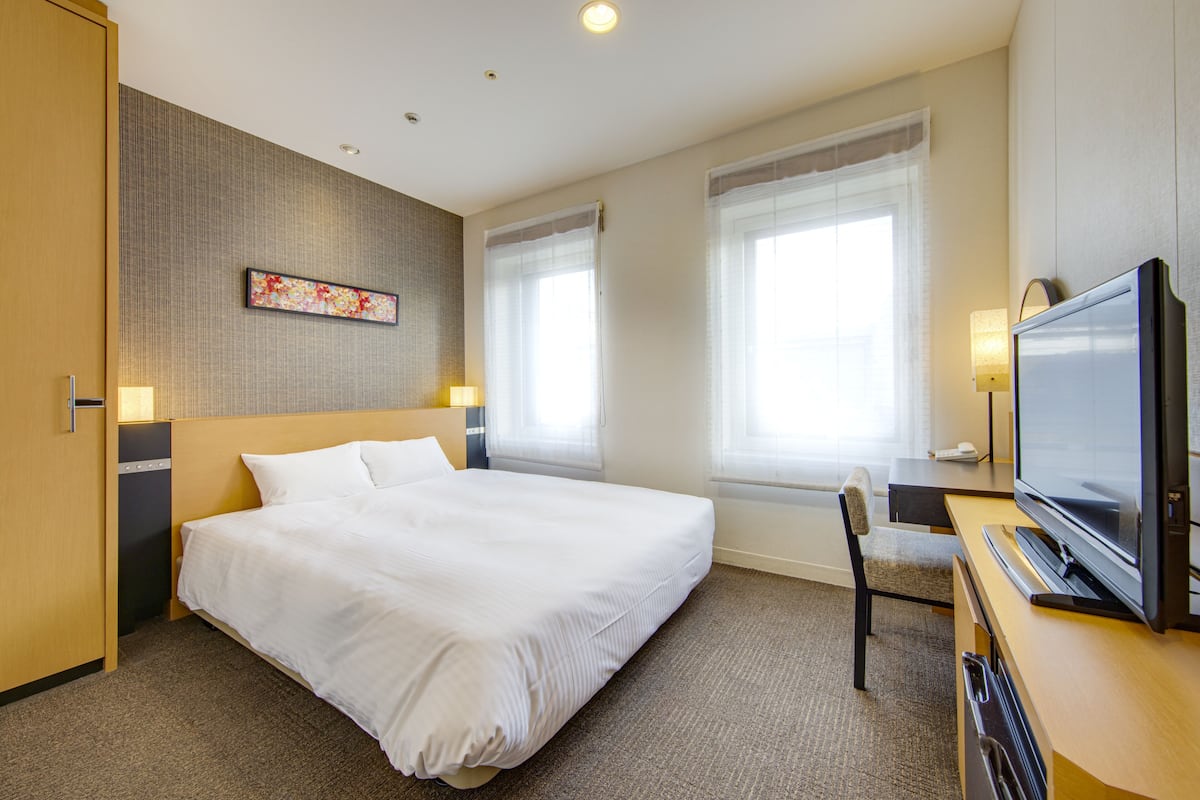 Quarto de hotel com uma cama grande, uma escrivaninha com televisão e duas janelas com cortinas.