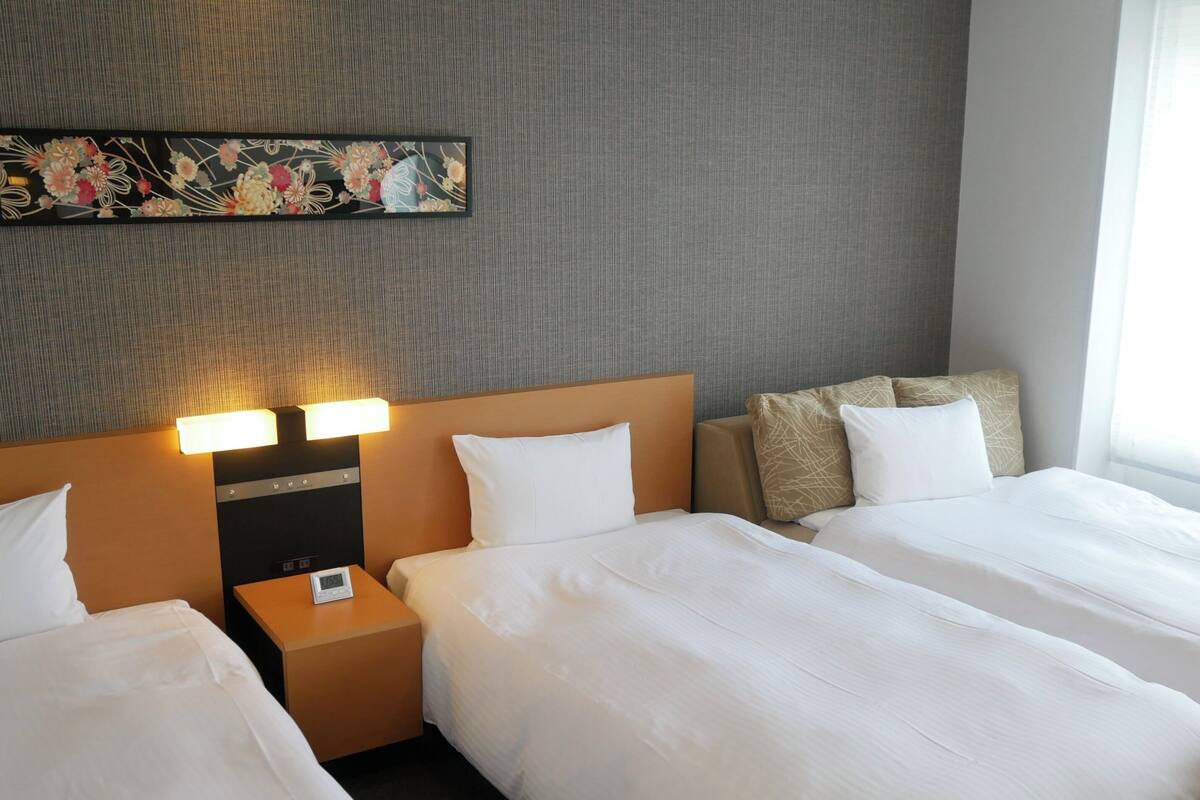 Quarto de hotel com duas camas, uma mesinha de cabeceira com abajur e um objeto decorativo na parede.