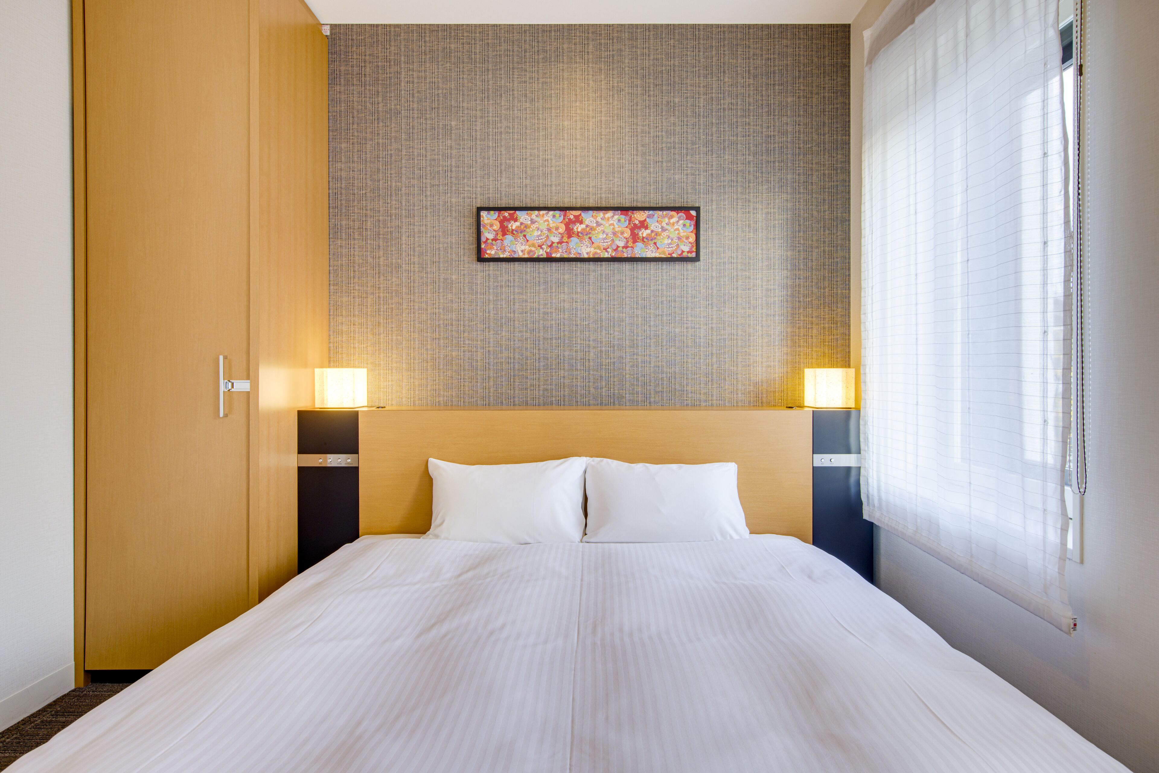 A hotel room with a large bed, two bedside lamps, a headboard, a wall-mounted artwork, and a window with blinds.