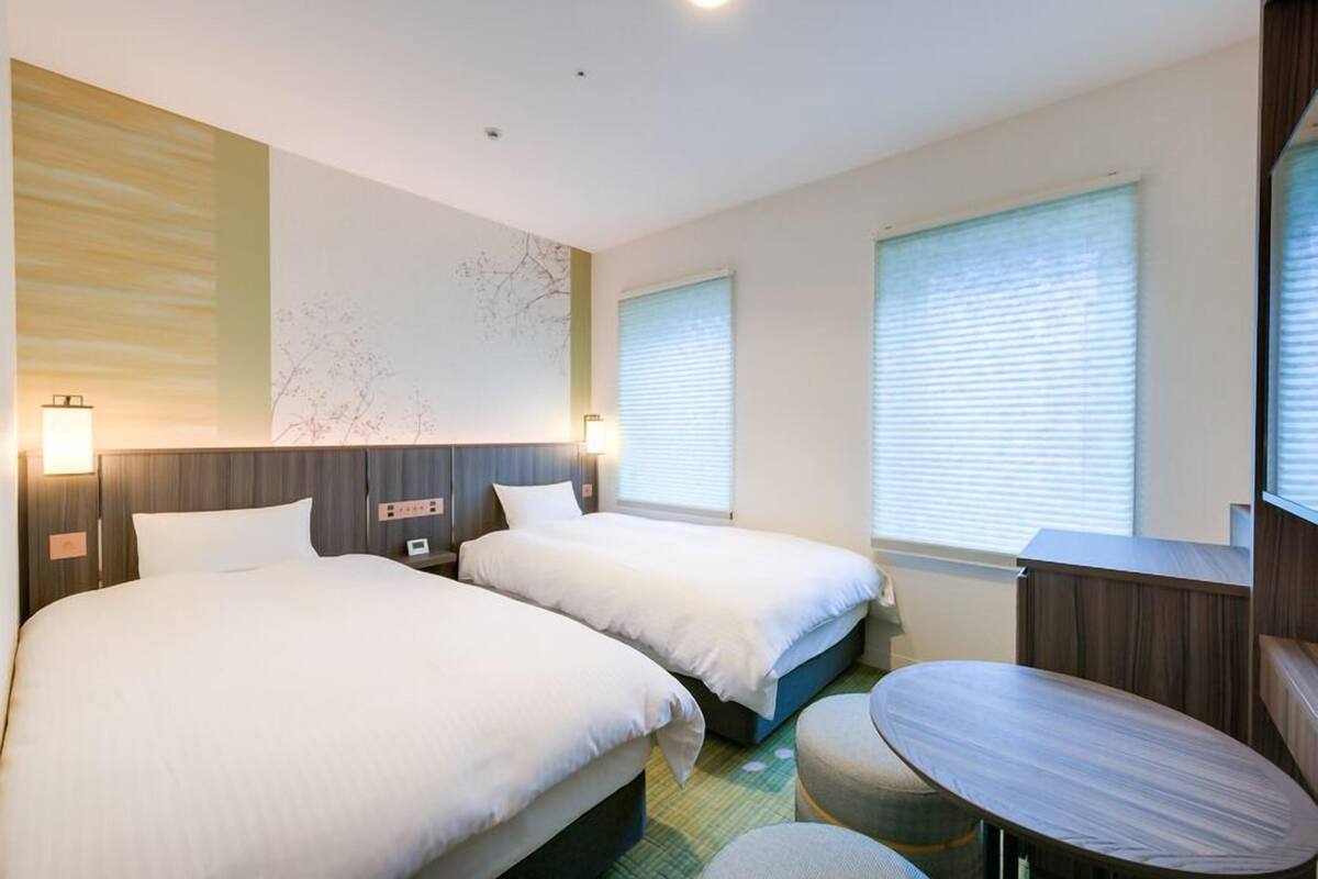 Quarto de hotel com duas camas, uma mesinha e uma luminária na parede.