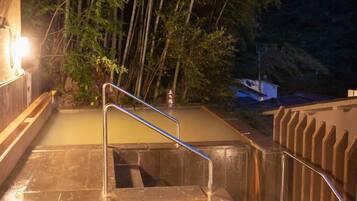A hot spring area with a metal staircase leading down to a pool of water, surrounded by trees and a building structure.