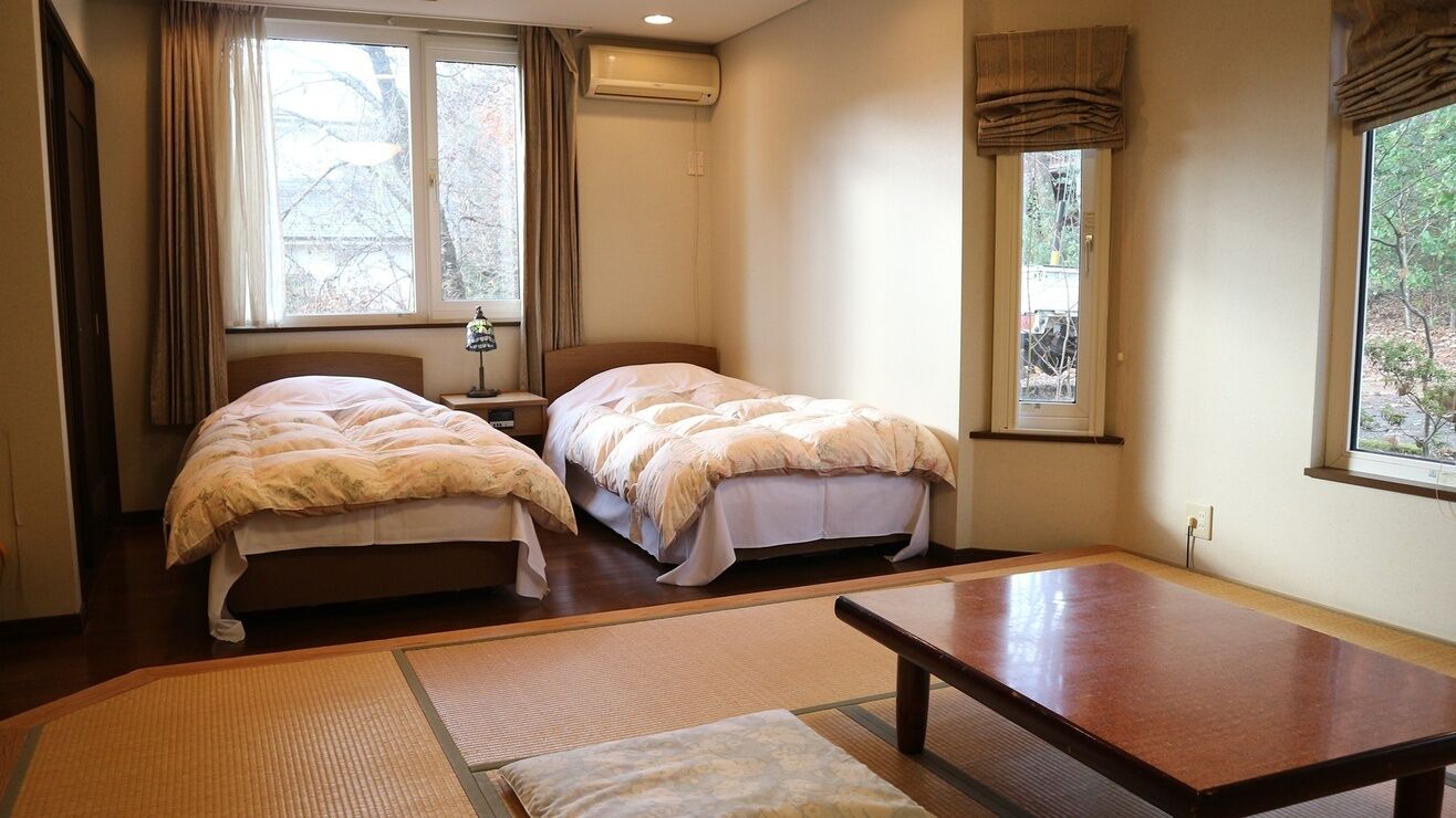 A room with two beds, a wooden table, and a view of trees through the window.
