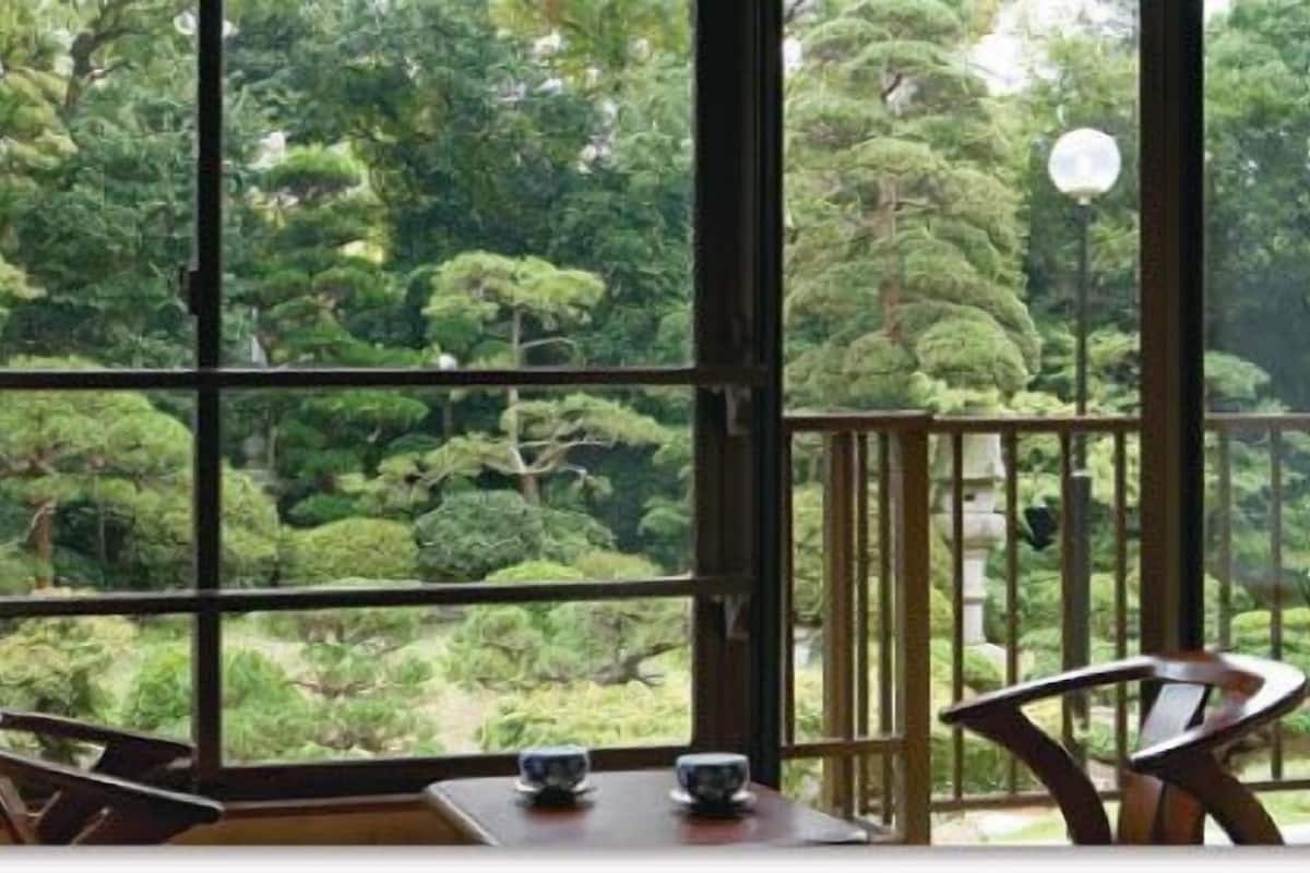 A room with a large window offering a view of a lush garden. There is a table with two chairs and a small table with cups.