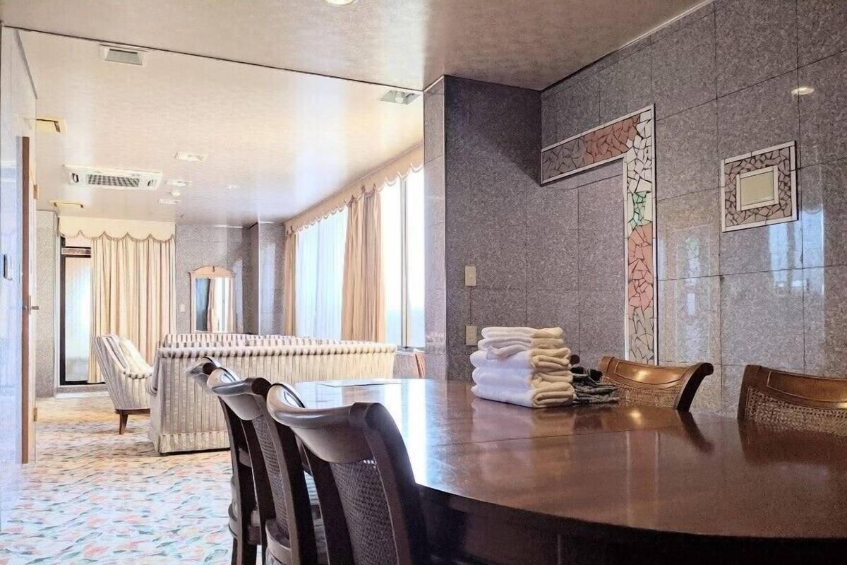 Habitación de hotel con una gran mesa redonda, varias sillas, un sofá y vistas de la ciudad a través de las ventanas.