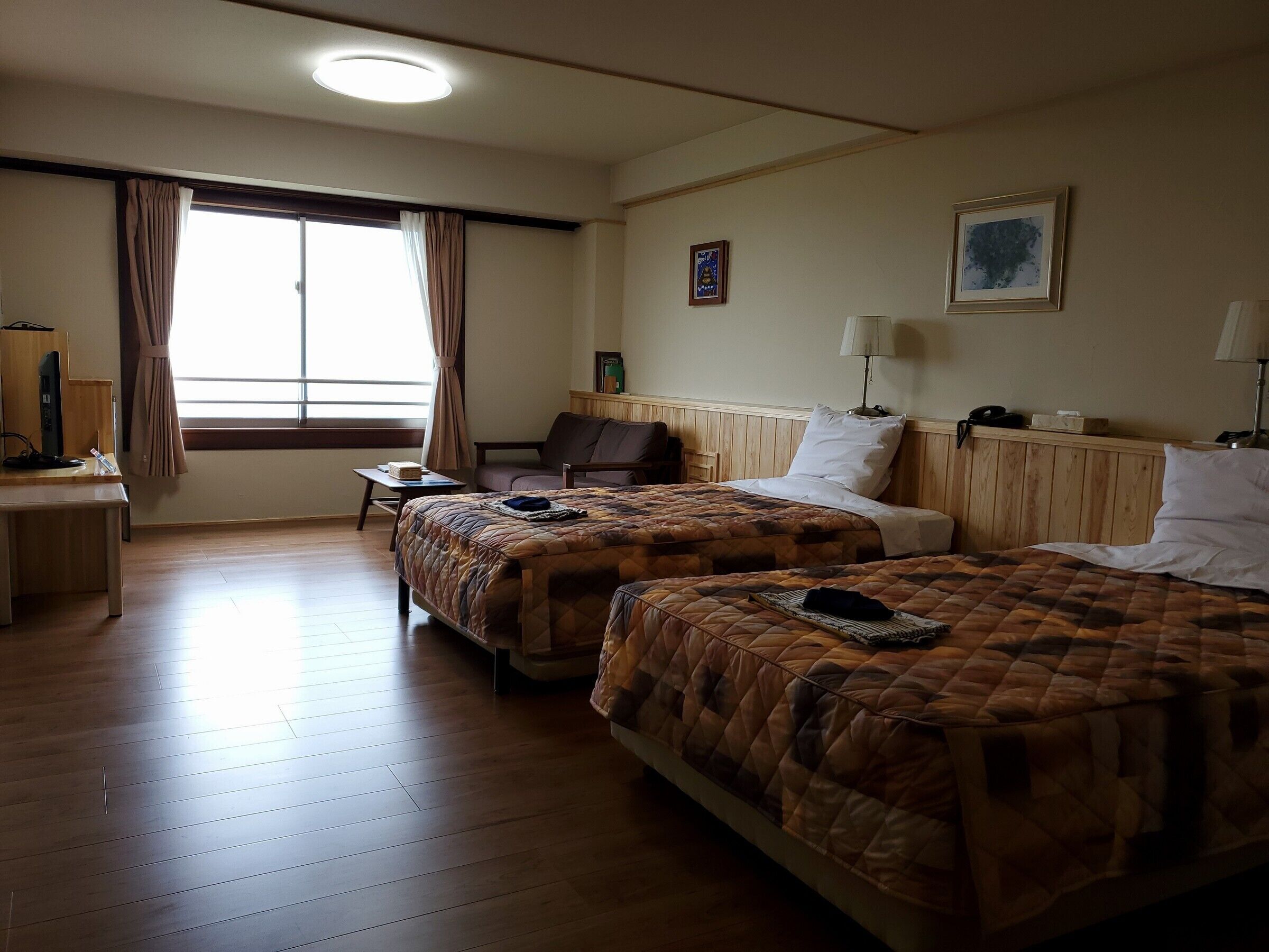 Een hotelkamer met twee bedden, een bureau, een stoel, een raam met gordijnen, een lamp en een ingelijst schilderij aan de muur.