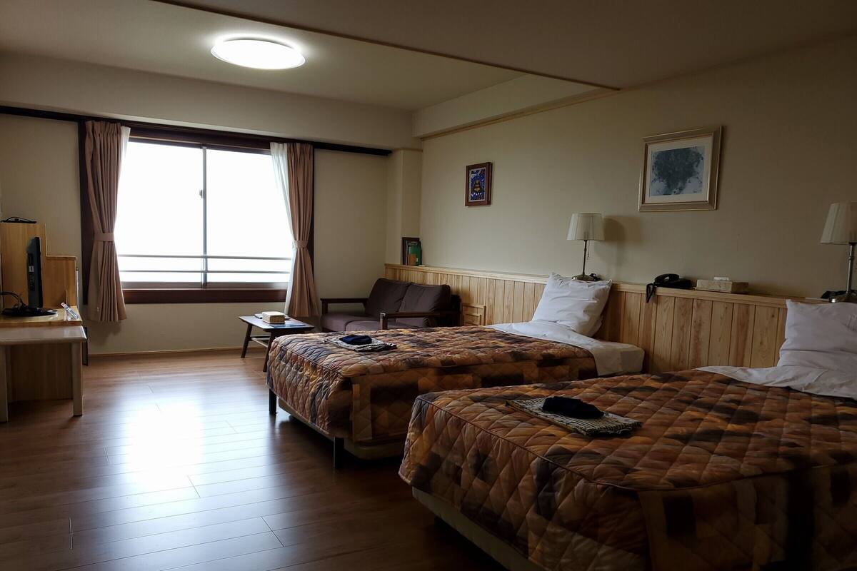 Une chambre d’hôtel avec deux lits, un bureau, une chaise, une fenêtre avec des rideaux, une lampe et un tableau encadré au mur.
