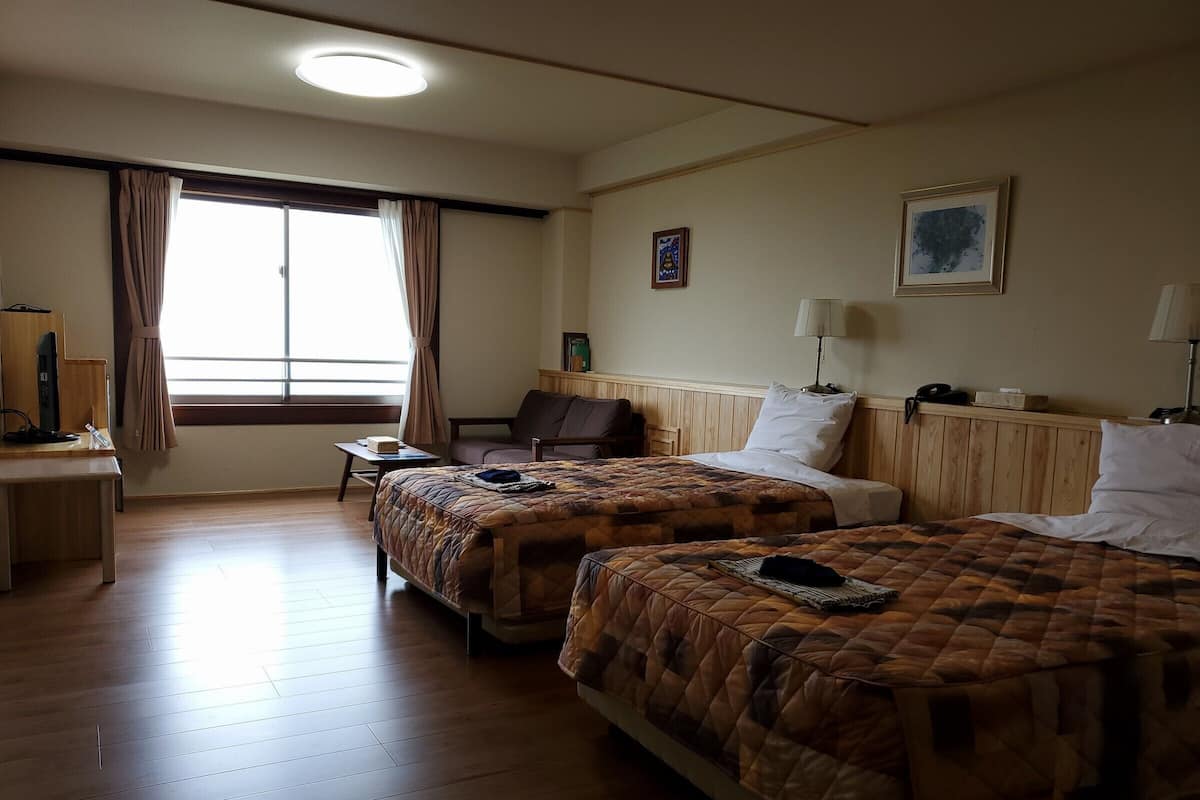 Een hotelkamer met twee bedden, een bureau, een stoel, een raam met gordijnen, een lamp en een ingelijst schilderij aan de muur.