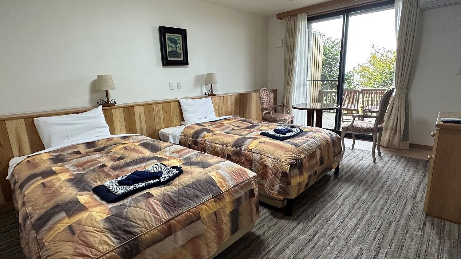 A hotel room with two beds, a small table, a chair, and a balcony with a view.