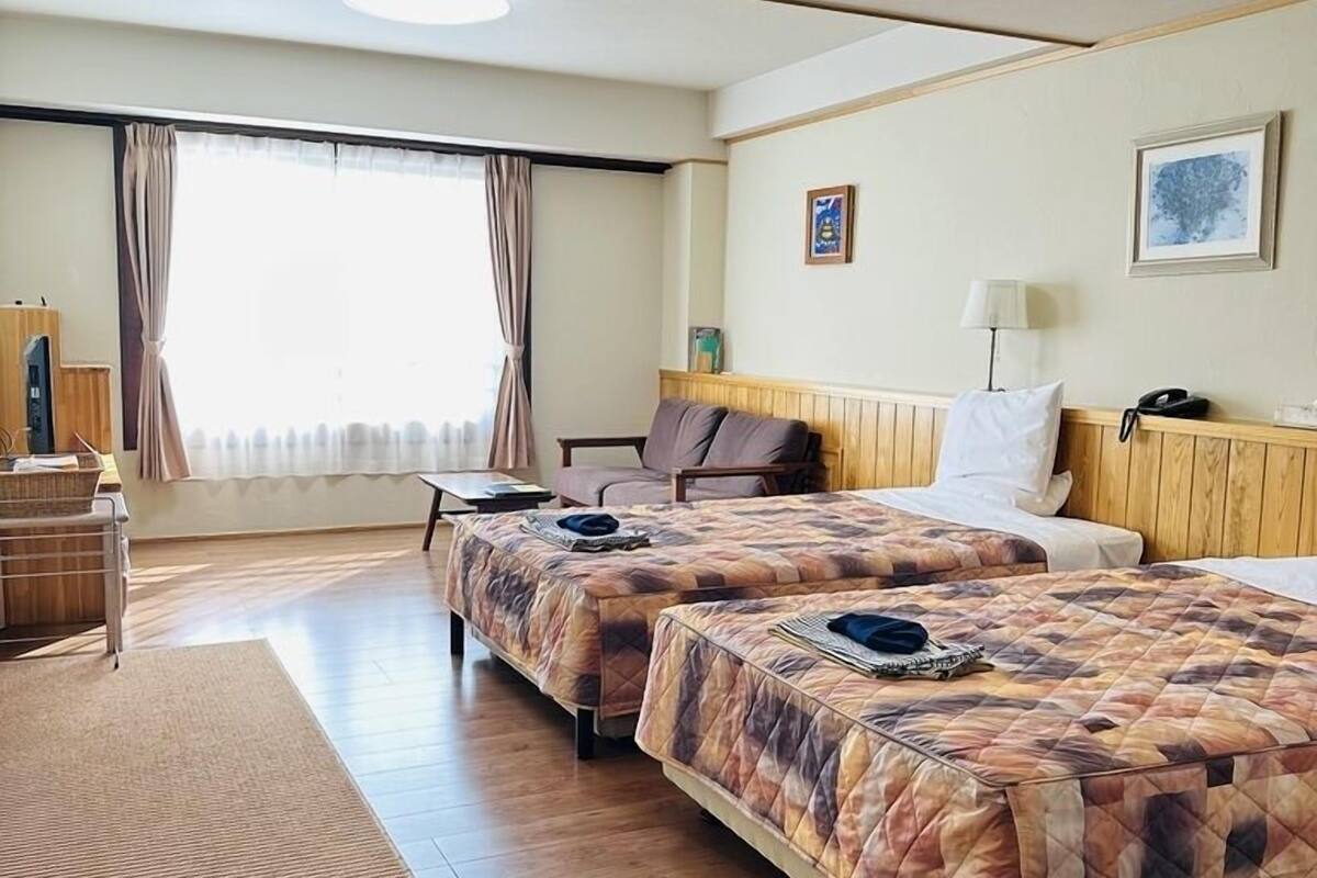 Une chambre d’hôtel avec deux lits, un bureau, une chaise, une fenêtre avec des rideaux et un mur orné d’un tableau.