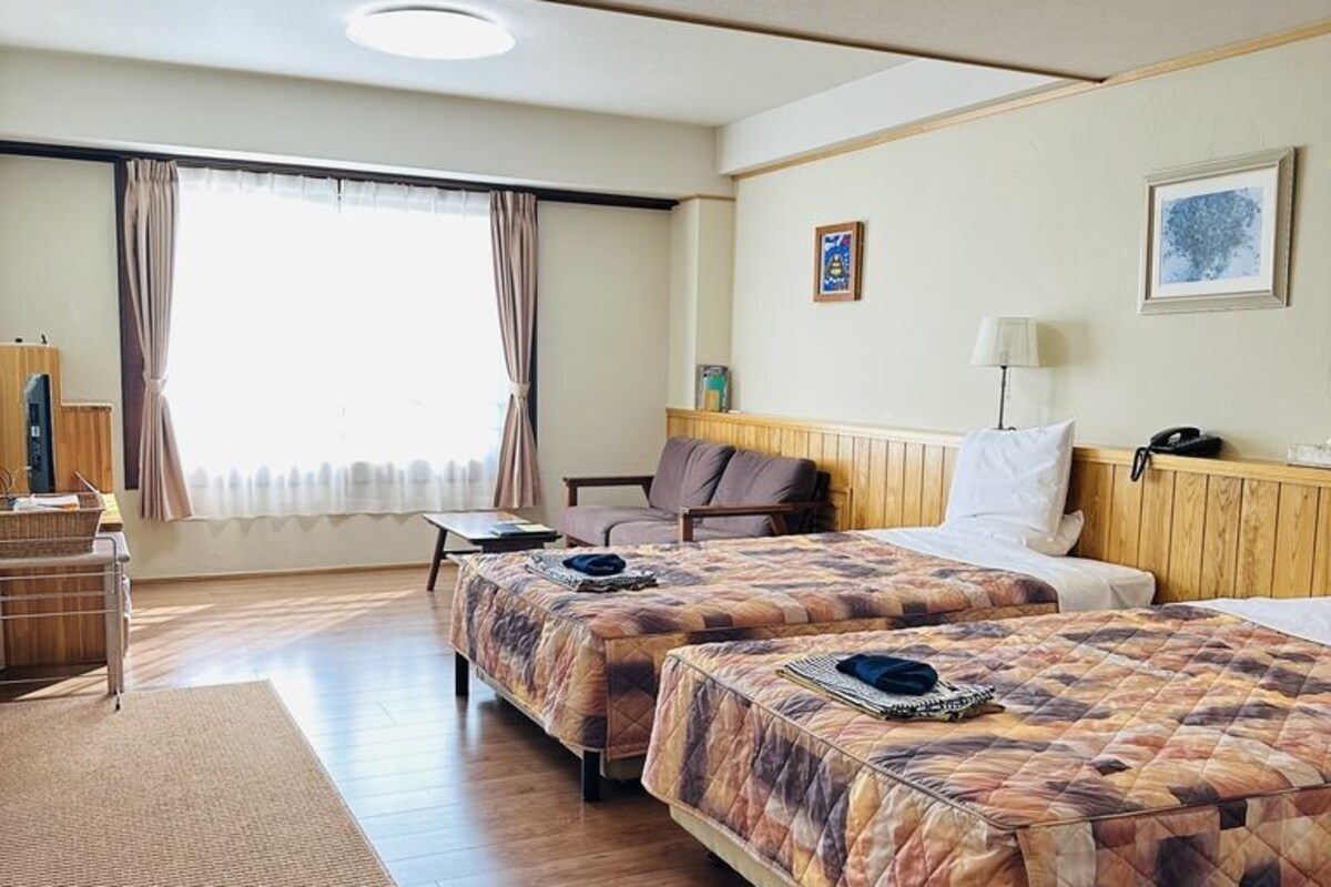 Une chambre d’hôtel avec deux lits, un bureau, une chaise, une fenêtre avec des rideaux et un mur orné d’un tableau.