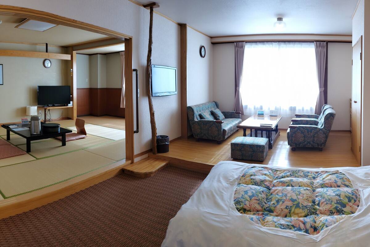 Une chambre d’hôtel avec un sol recouvert de tatamis, un lit avec une literie à motifs floraux, un canapé, une télévision et un bureau avec une lampe.